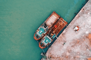 Aerial view of two large cargo ships docked at a port, tied to the pier, with turquoise water on the left and a gravel dockyard on the right. Represents global shipping operations and port congestion.